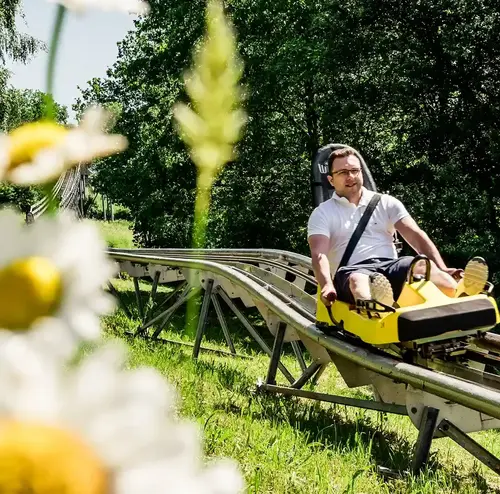 Verschwommene Blumen im Vordergrund, im Hintergrund ein Mann auf einem gelben Rodelschlitten der Rodelbahn in Gutach.