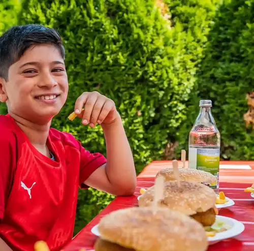 Ein Kind sitzt lachend im Imbissbereich der Sommerrodelbahn Gutach vor einem Teller mit Pommes und Hamburger.