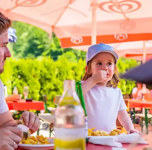 Ein Kind und ein Mann sitzen unter Sonnenschirmen im Imbissbereich der Sommerrodelbahn Gutach und essen Pommes.