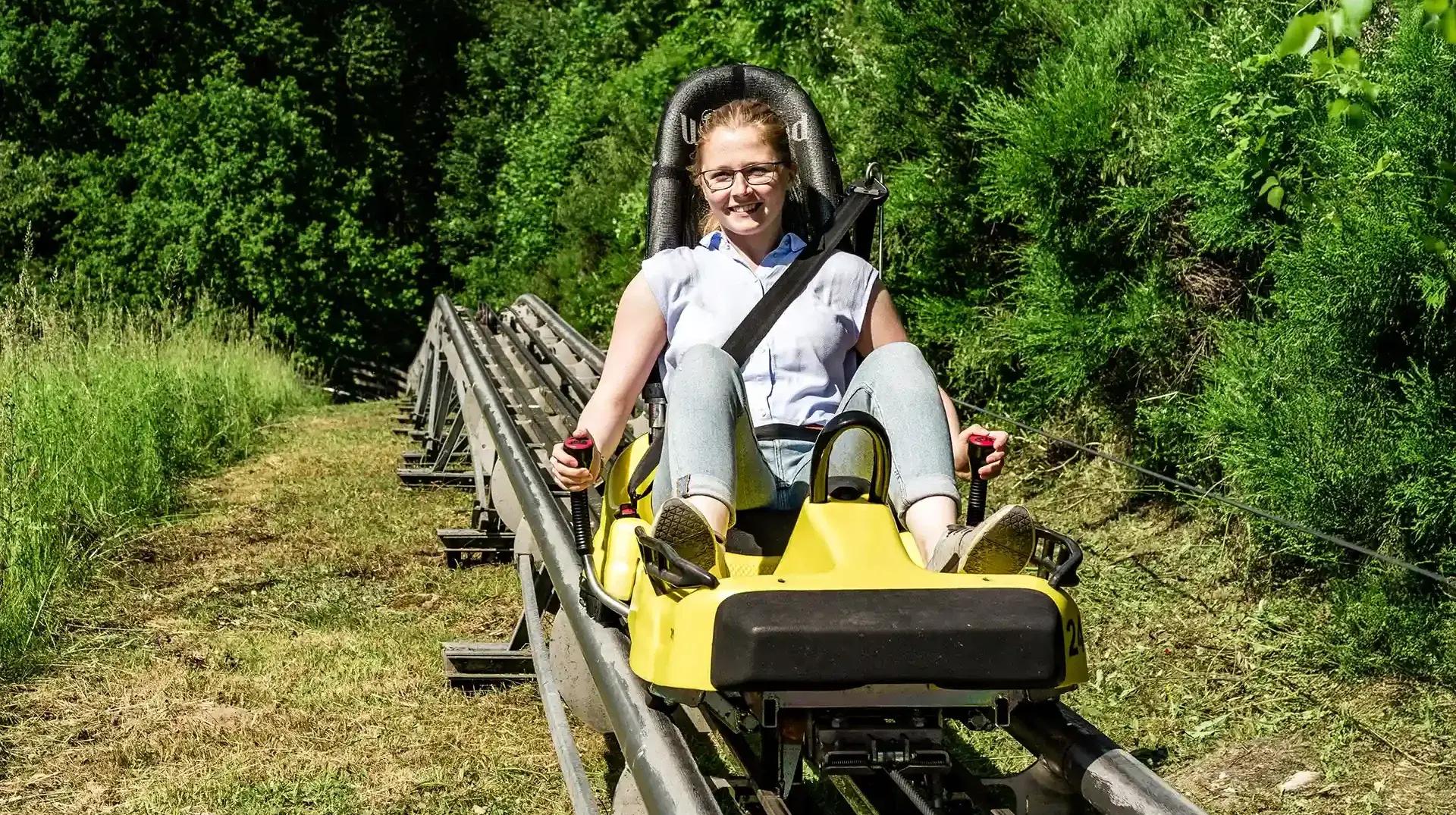 Eine Frau hält lächelnd die Bremshebel eines gelben Alpine Coaster Schlittens, im Hintergrund grüne Vegetation.