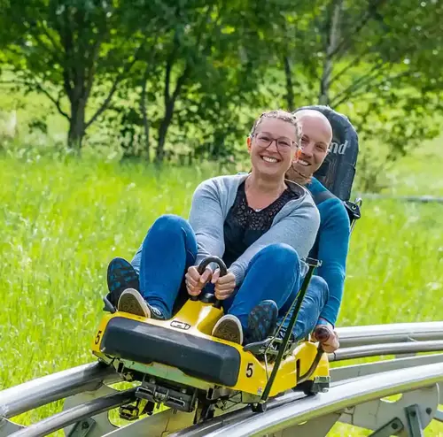 Ein Mann und eine Frau fahren lachend auf der Sommerrodelbahn in Gutach durch eine Wiesenlandschaft.