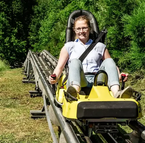 Eine Frau sitzt lächelnd in einem gelben Alpine Coaster Schlitten, im Hintergrund Bäume und Büsche.